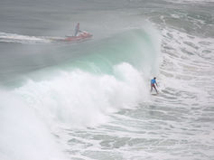 A surfer rides a towering wave while a jet ski follows close behind, emphasizing the immense scale and energy of Nazaré’s waters
