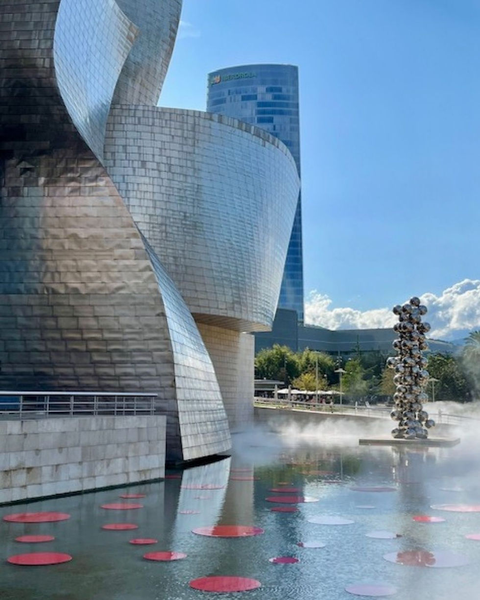 Modern building with metallic curves by a pond with red circles, mist, and a shiny sculpture. Blue sky and a glass tower in the background.