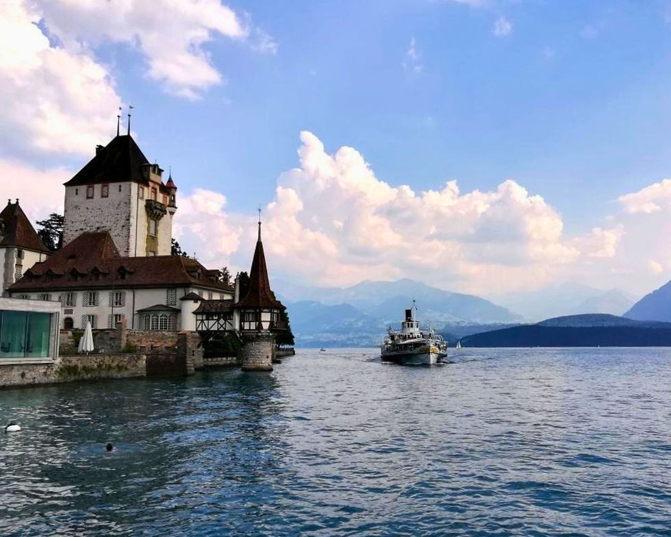 Historic castle by a lake with a boat approaching, set against a backdrop of mountains and a partly cloudy blue sky. Serene and picturesque.