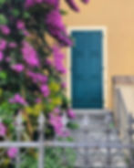 Teal shuttered door on a yellow wall, framed by vivid purple bougainvillea and a wrought-iron fence, calm garden entrance