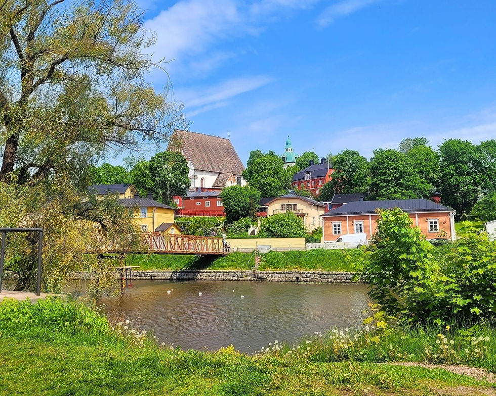 Quaint village scene with colorful houses by a river, lush greenery, a wooden bridge, and a distant church under a bright blue sky.