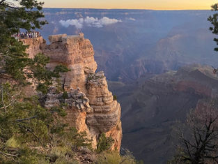 People stand on a rocky cliff at the Grand Canyon during sunset. Trees frame the scene, and the sky is a gradient of blue and orange.