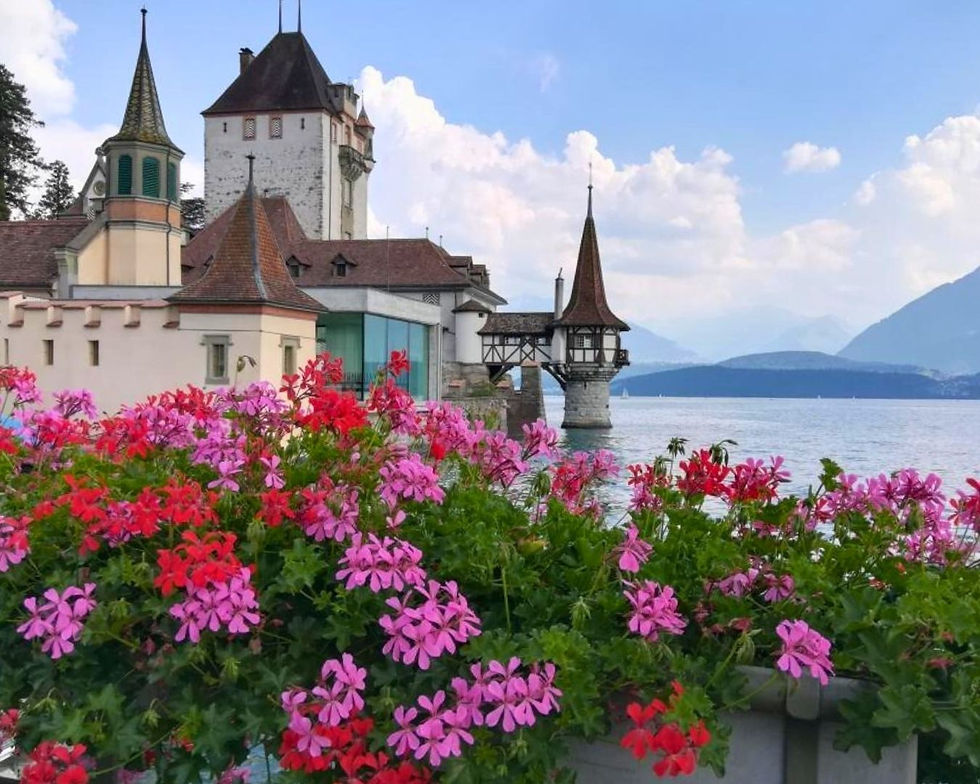 Castle by a serene lake, framed by pink and red flowers, with a mountainous backdrop under a partly cloudy sky, creating a peaceful scene.