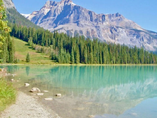 Turquoise lake with rocky shore reflecting a forest and majestic mountains under a clear blue sky. Peaceful and serene setting.