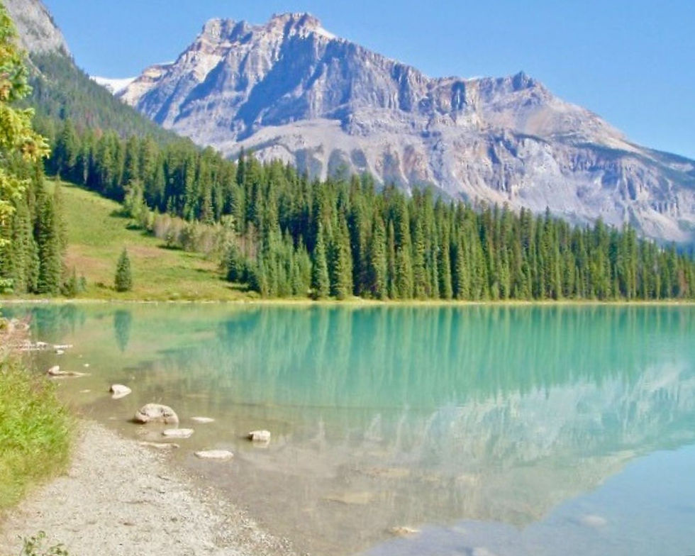 Turquoise lake with rocky shore reflecting a forest and majestic mountains under a clear blue sky. Peaceful and serene setting.