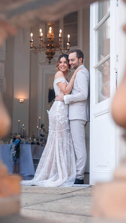Ein Brautpaar steht am Eingang eines Schlosses mit einen Kronleuchter hinter ihnen und blickt glücklich nach draußen, fotografiert von Raluca Wassmus in Villa Eugenia