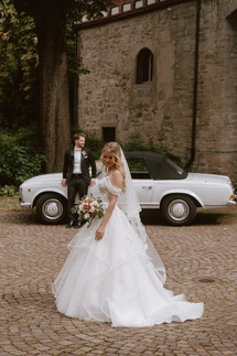 Braut im eleganten Hochzeitskleid vor einem weißen Oldtimer, Bräutigam steht im Hintergrund – stilvoller Fine-Art-Moment.
