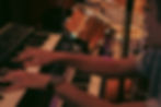 A set of two keyboards, and a pianists arms and blurred side profile, with a sparkly drum kit in the background, a warm photo of a Wombat Jazz Club event in a basement on Bold Street, Liverpool.