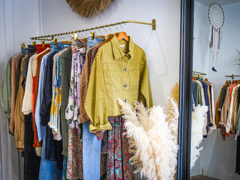 Clothing rack with colorful garments; a khaki jacket is prominent. Mirrors, pampas grass, and a dreamcatcher in the cozy store setting.