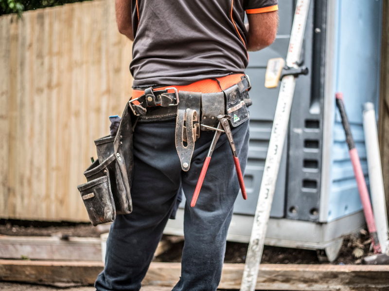 Worker with tool belt stands near a construction site, wearing a gray and orange shirt. Tools include pliers. Wooden fence in background.