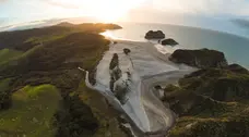 A strip of sandy beach among grassy hills, looks out to sea as the sun sets.