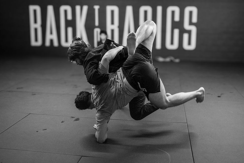 Two men grapple (doing Boston jiu jitsu) on a mat in intense jiu-jitsu training. "Back to Basics" is visible in the blurred background.