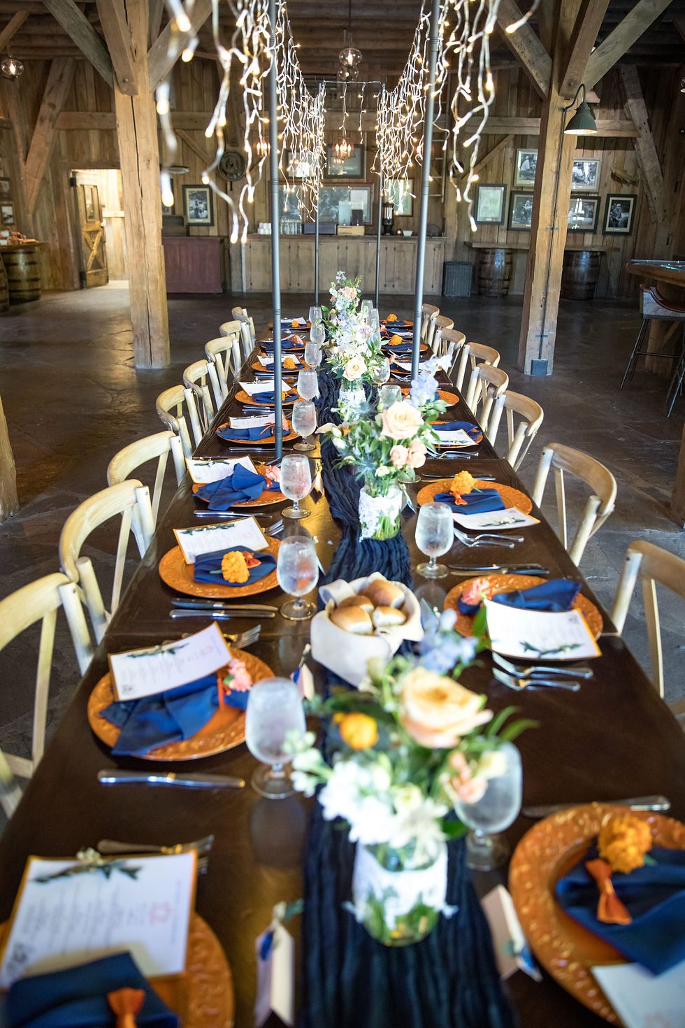 A long wedding reception table, set with chargers, menus, napkins, floral centerpieces, and lights hanging overhead.