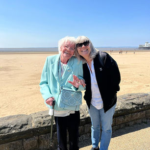 Resident and carer smiling in front of a bright sunny beach.