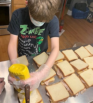 An adolescent is preparing a large number of sandwhich, and is shown wearing a facemaks and plastic gloves as he squeezes a mustard bottle.