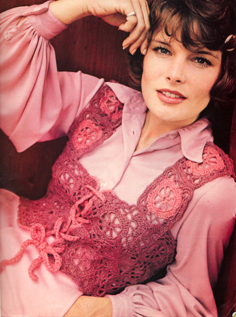 A woman poses wearing a pink collared dress with a pink crocheted vest over top. 