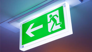 Green illuminated exit sign with a running figure and left arrow, mounted on a ceiling. Bright and clear guidance for emergencies.