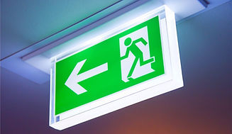 Green illuminated exit sign with a running figure and left arrow, mounted on a ceiling. Bright and clear guidance for emergencies.