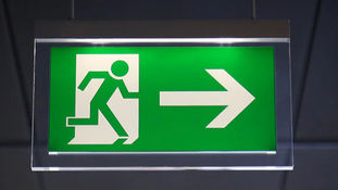 Green exit sign showing a figure running through a door with a right arrow, suspended against a dark, blurred ceiling background.