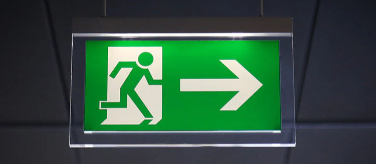 Green exit sign showing a figure running through a door with a right arrow, suspended against a dark, blurred ceiling background.