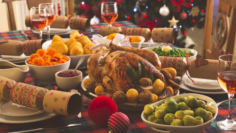 Festive Christmas dinner table with roast turkey, veggies, wine, and crackers on plaid cloth. Decorated tree in the background. Cozy mood.