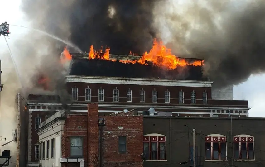 A large brick building on fire with flames and smoke rising. A firefighter sprays water from a ladder to douse the flames.