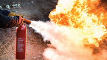 A person uses a red fire extinguisher to put out bright orange flames on a rough surface. White foam sprays onto the fire.