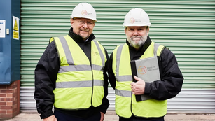 Two men in safety vests and helmets stand smiling in front of a green industrial door. One holds a folder labeled "FS Industrial Fire Safety Limited."