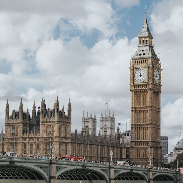 Clock tower and parliament building under a cloudy sky. Tourists and red buses on the bridge create a lively urban scene.