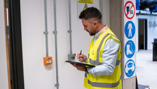 Man in a yellow safety vest writes on a clipboard in an industrial setting. Safety signs on the wall. White and grey background.