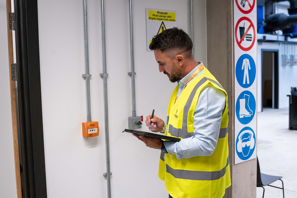 Man in a yellow safety vest writes on a clipboard in an industrial setting. Safety signs on the wall. White and grey background.