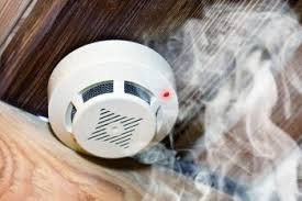 A white smoke detector on a wooden ceiling releases smoke, indicating activation. A red light is visible, creating a sense of urgency.