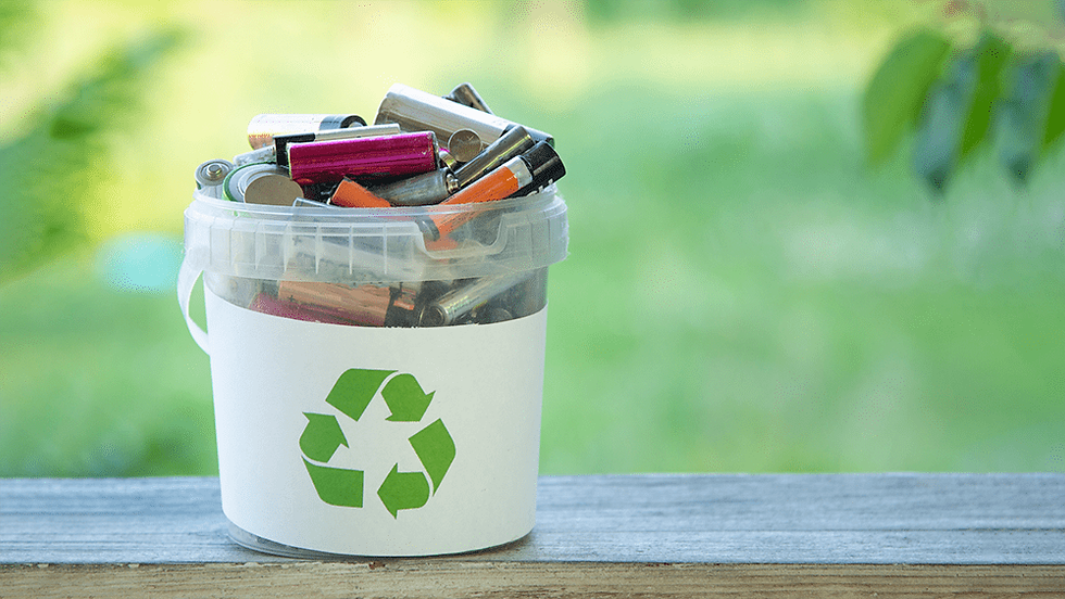 A small bucket filled with various batteries, labeled with a green recycling symbol, sits on a wooden ledge against a blurred green background.