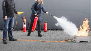 Two people demonstrate fire extinguisher use outdoors. A woman sprays foam on a controlled flame; red extinguishers and cables are visible.