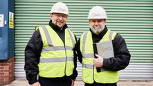 Two men in hard hats and reflective vests stand smiling in front of a green shutter. One holds a folder labeled "FS Fire Safety Limited."