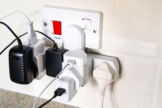 Multiple plugs and adapters are crowded into a white wall socket. A red switch is visible. The background has light-colored tiles.