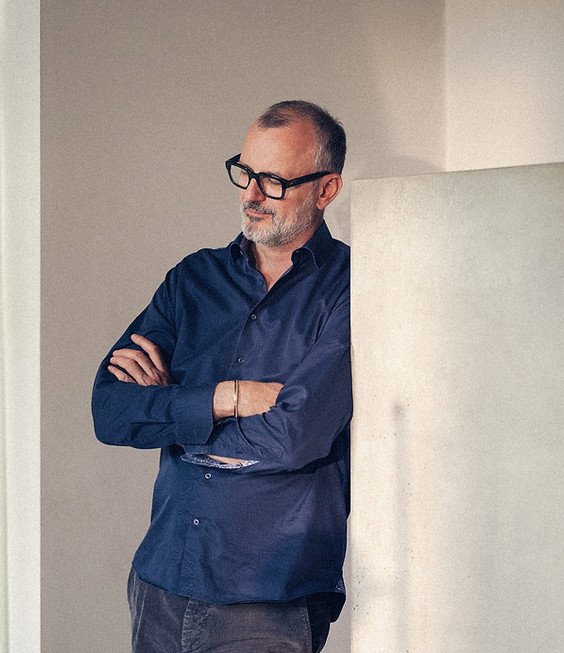 Mann mit Brille und blauem Hemd lehnt mit verschränkten Armen an einer Wand in einem hellen Raum