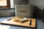 Sliced bread arranged on a wooden cutting board with a butter knife sits in a kitchen setting, beside a bread bin labelled "B