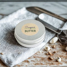 A small tin of butter marked "Coopers Butter" sits on a textured white cloth with two silverware utensils, with some seeds scattered around. The image has a rustic, homey vibe, with a focus on natural elements.