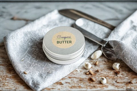 A round container of butter sits on a patterned napkin next to a silver butter spreader, with a few decorative seeds scattere