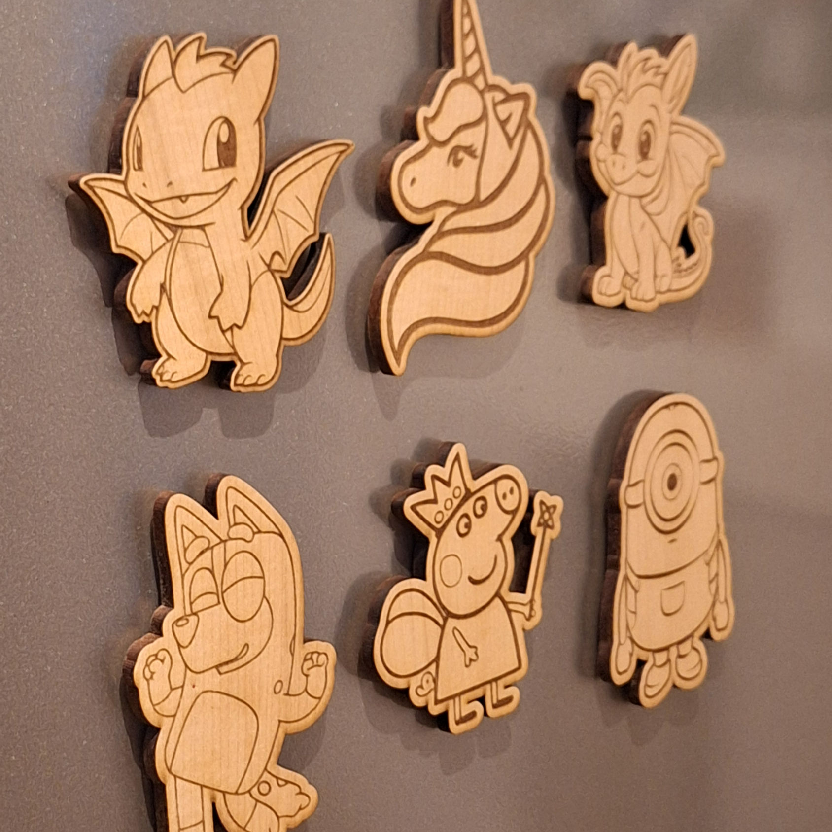 Six wooden magnets, shaped and laser-cut like various cartoon characters, are attached to a gray metal surface.  The characte