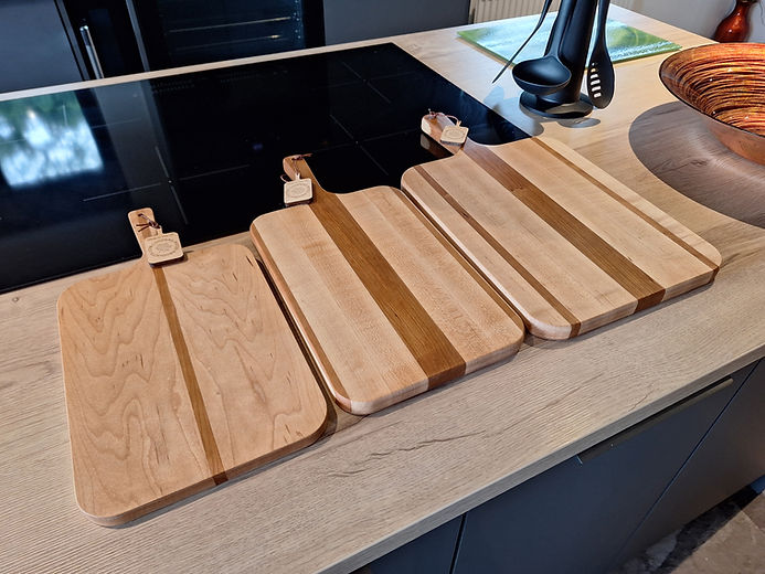 Three handcrafted maple wooden cutting boards with differing stripe patterns are artfully displayed against the backdrop of a modern kitchen countertop and sleek black stovetop. Each board features a light-toned wood with darker wood accents.
