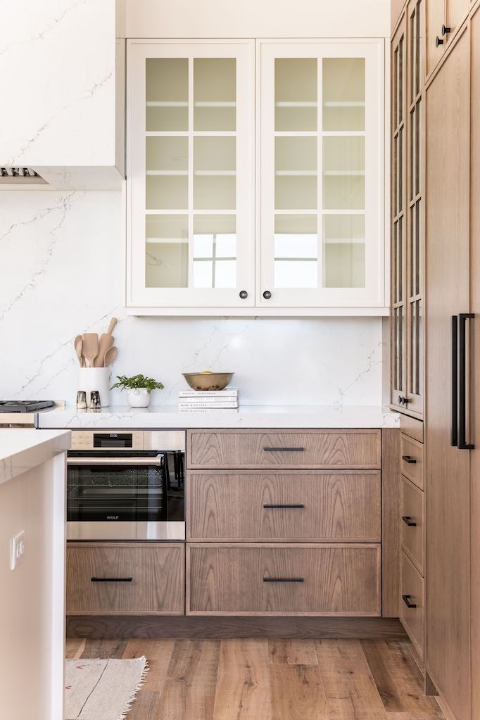 Modern kitchen with wood cabinets, marble countertops, and glass-paneled cupboards. Neutral tones, wooden utensils, and potted plant add warmth.