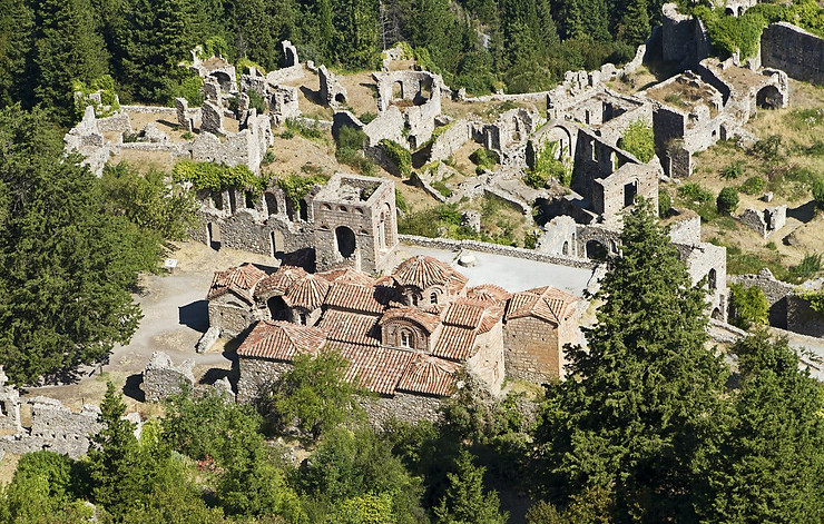 Luftaufnahme der byzantinischen Ruinen von Mystras mit Steingebäuden und Bögen, umgeben von Grün, und den charakteristischen roten Ziegeldächern eines Klosters in der Mitte.