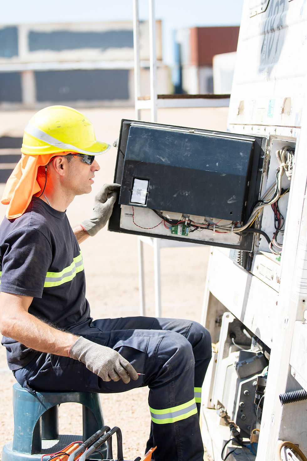 Trabajador con casco amarillo y guantes manipula un panel eléctrico al aire libre. Lleva uniforme azul con reflejantes. Fondo con contenedores.