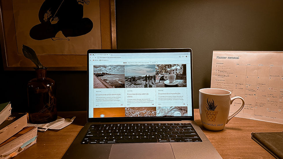Computador aberto na página do blog sobre uma mesa de trabalho. À direita, um planner mensal e uma xícara de café. À esquerda, livros empilhados e papéis sob uma garrafa de vidro com água. Dentro do copo, uma planta criando raízes, sugerindo crescimento lento e contínuo em um ambiente cotidiano.
