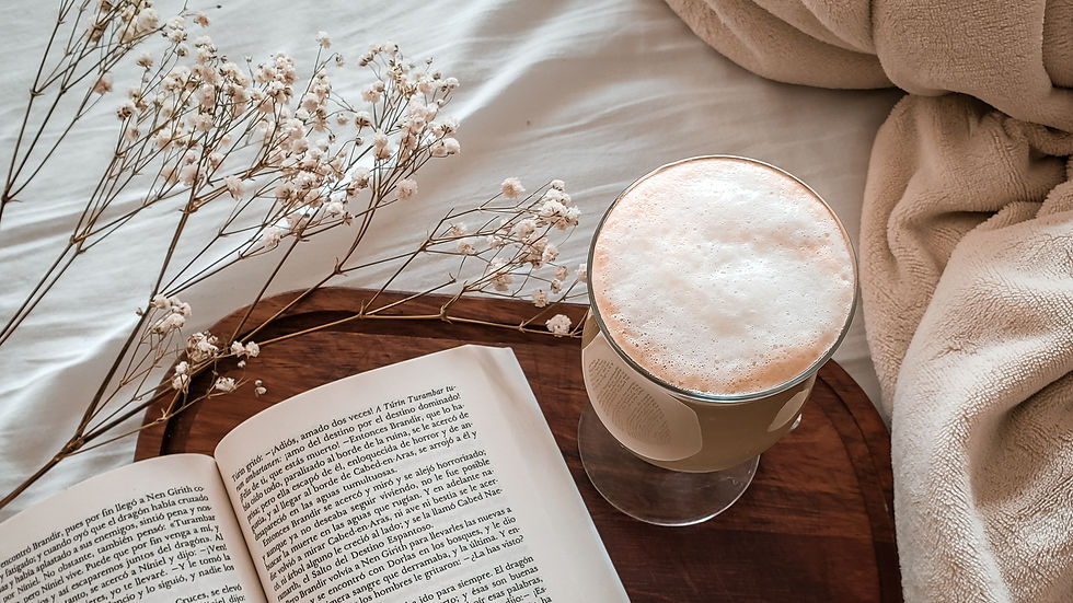 Uma xícara de cappuccino e um livro descansam sobre uma bandeja na cama, cercados por um cobertor macio e flores secas representando momentos simples e reais do cotidiano.