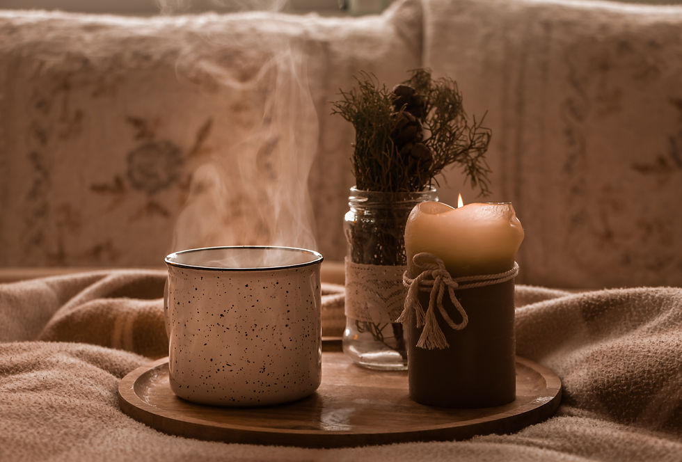 Uma xícara de café esfumaçante, uma vela acessa, um ramo de folhas de pinheiro, dentro de um vaso de vidro, como um adorno decorativo, sobre uma bandeja de madeira, em cima da cama, ao fundo travesseiros e luz suave.