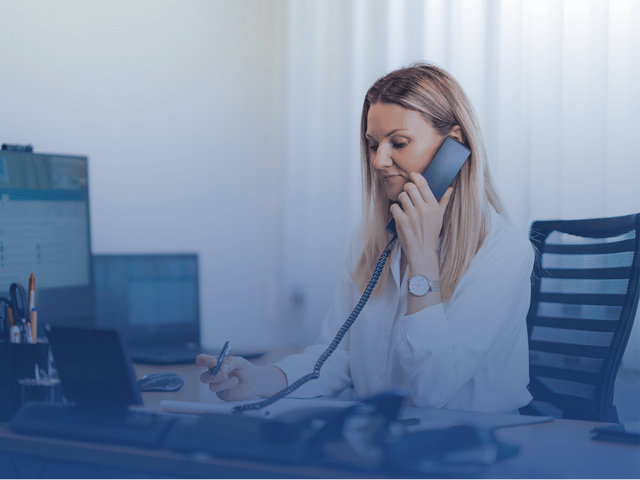Frau telefoniert am Schreibtisch im Büro mit einem Festnetztelefon und benutzt dabei einen Laptop.