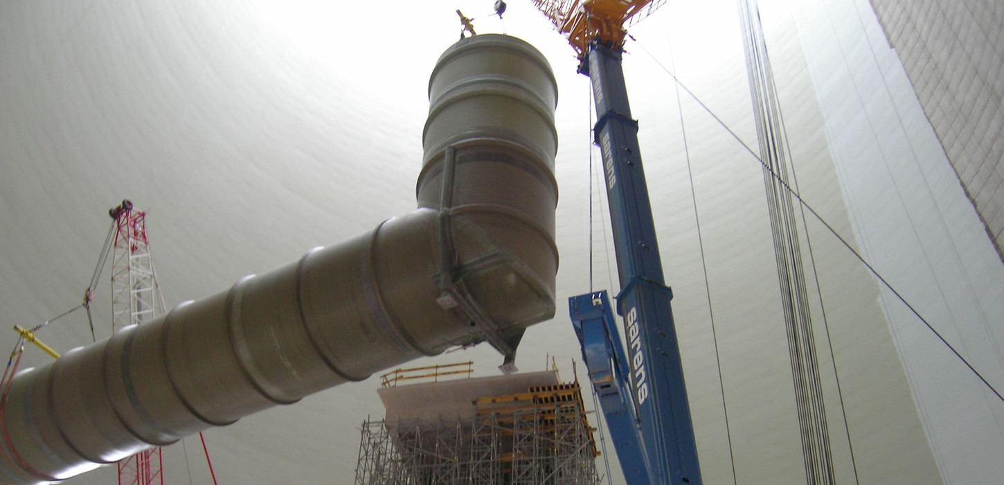 Ein schwerer Kran hebt ein großes Industrierohr innerhalb einer massiven Kuppelkonstruktion an.